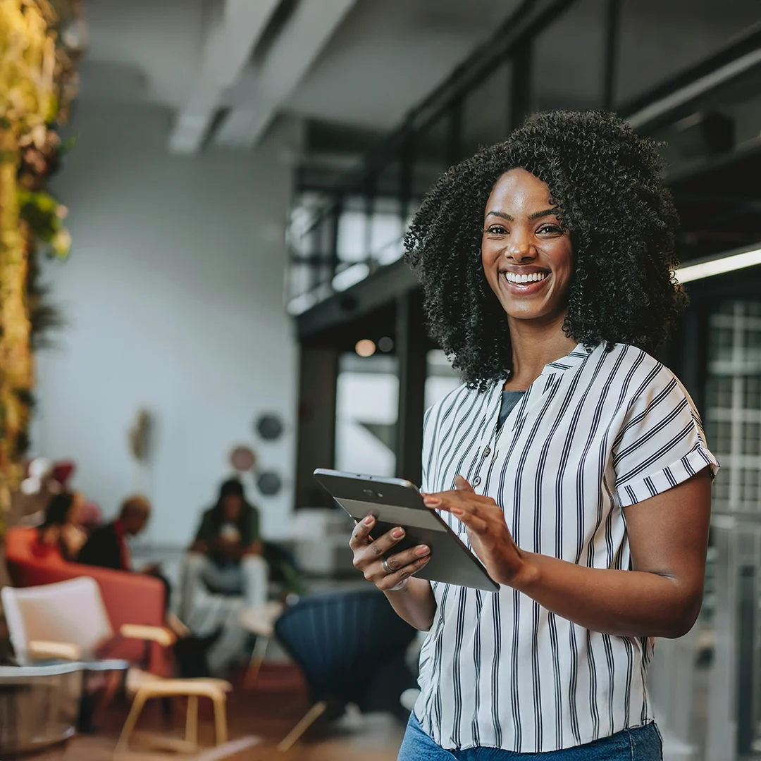 A smiling professional holding a tablet while standing in a modern office lounge with seating and coworkers in the background.