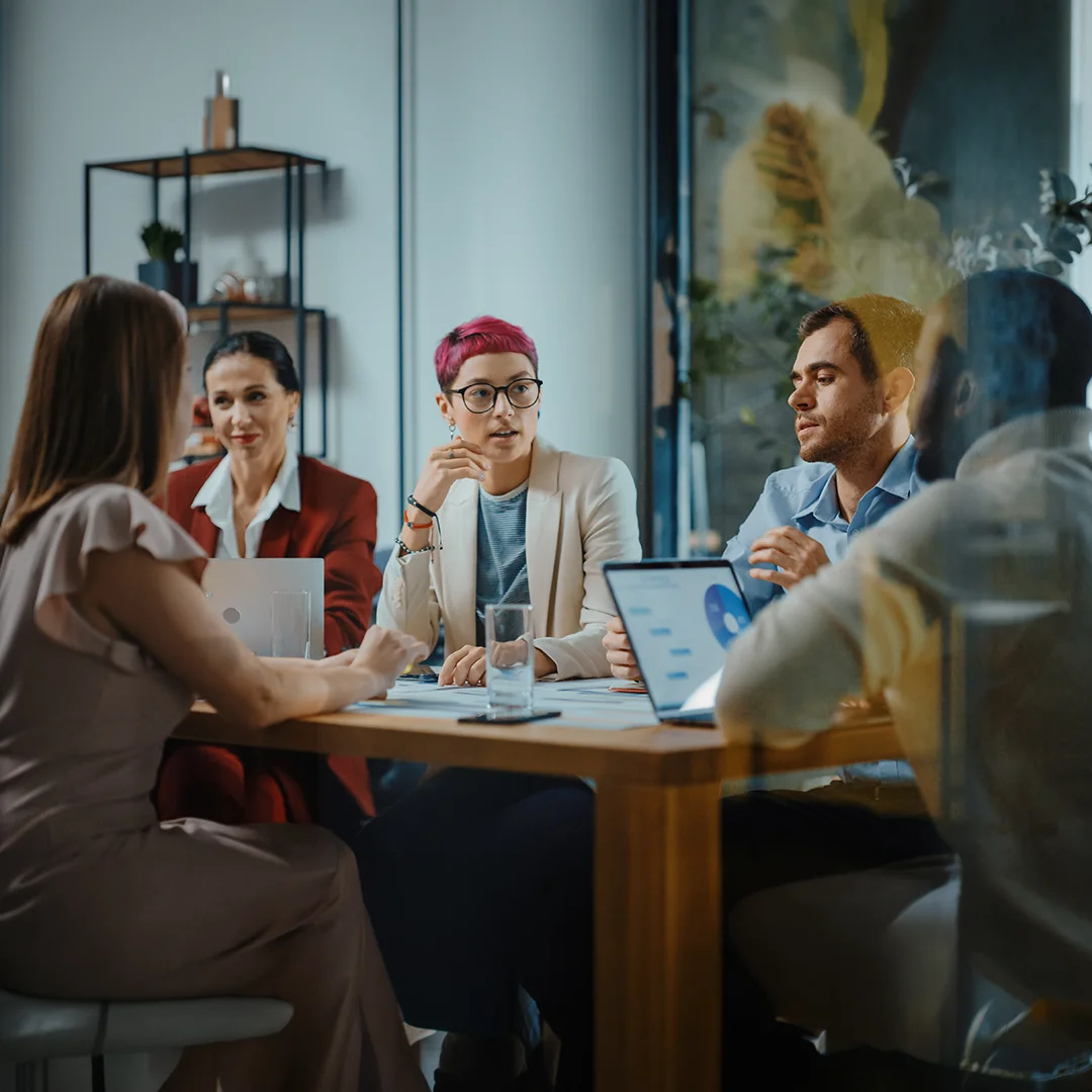 A group of professionals exchanges ideas around a conference table with laptops and printed materials.